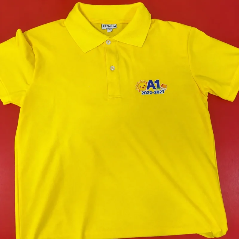 56 áo lớp polo A1 màu vàng – Vải cá sấu 80% cotton – Mặt trước áo polo A1 vàng với hình ngực trái và niên khóa 2022–2027. – 1