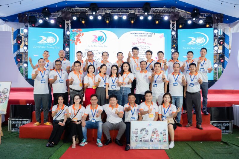 314 áo họp khóa K29 THPT Giao Thủy B do VIE Uniform sản xuất – Nhóm cựu học sinh K29 THPT Giao Thủy B chụp ảnh trong áo polo trắng phối cam trước sân khấu hội khóa. – 2